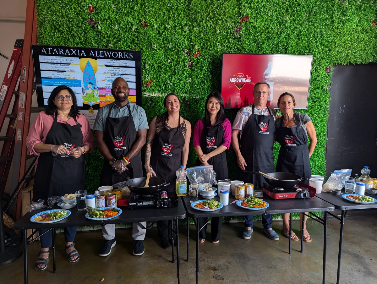 Participants in an Authentic Pad Thai cooking class stand at tables with ingredients, smiling against a green backdrop.