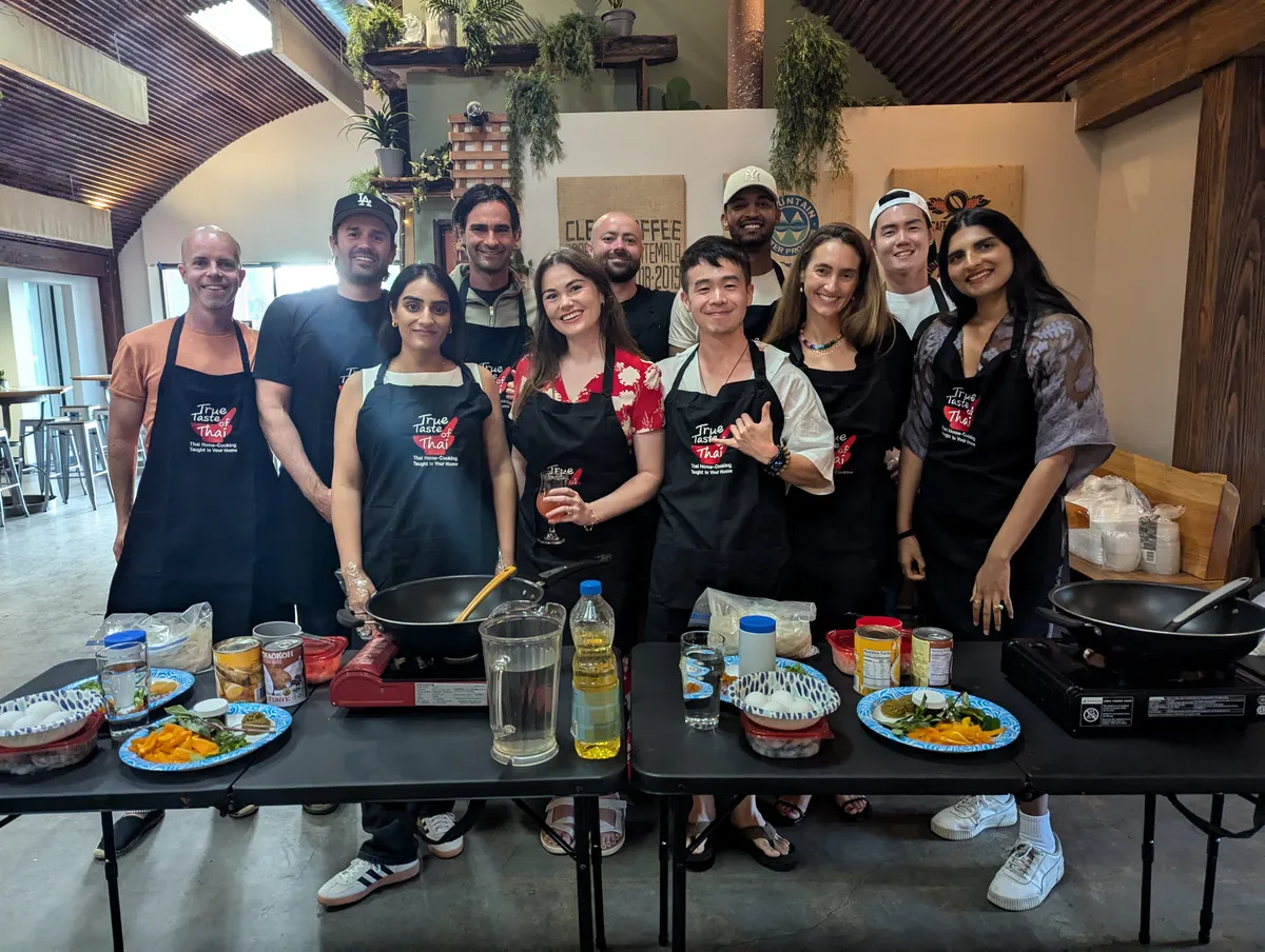 Group of participants wearing aprons in a cooking class, smiling around tables with ingredients for Pad Thai.
