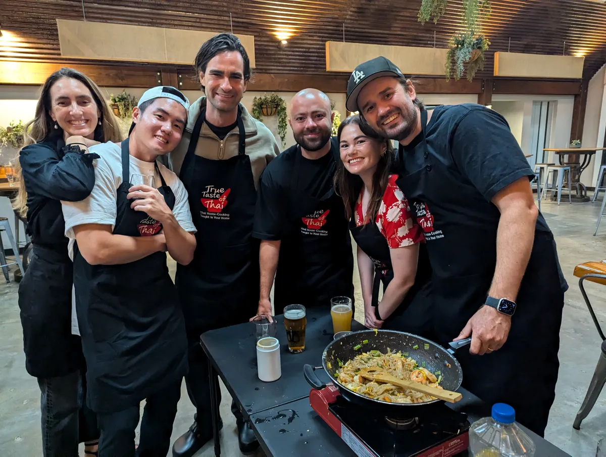 A group of six people in aprons smiles together by a cooking station featuring a wok of Pad Thai.