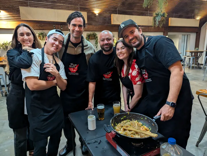 A group of six people in aprons smiles together by a cooking station featuring a wok of Pad Thai.