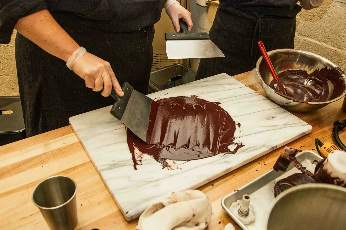 Hands using metal spatulas to spread dark chocolate on a marble surface during a Bean-to-Bar chocolate class.