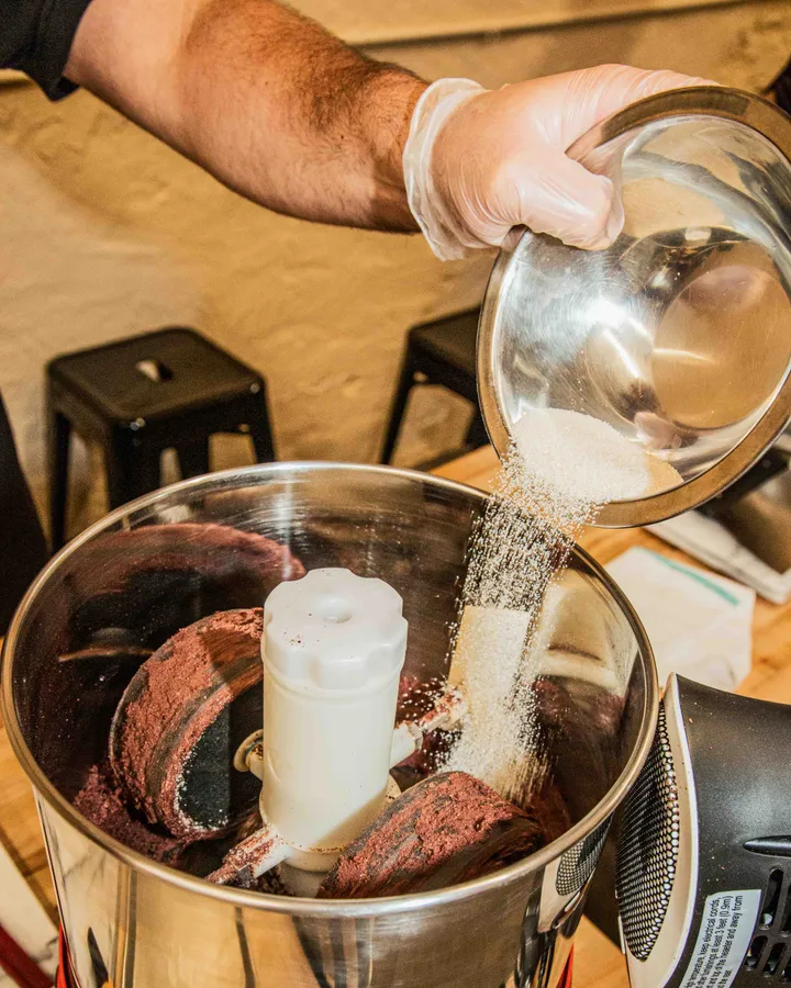 A hand pours sugar into a blender with chocolate mix during a Bean-to-Bar Chocolate Class in NYC.