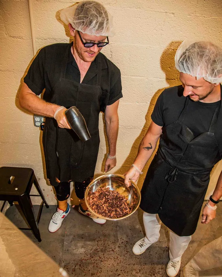 Two participants in aprons and hairnets work together in a chocolate-making class, mixing ingredients in a bowl.