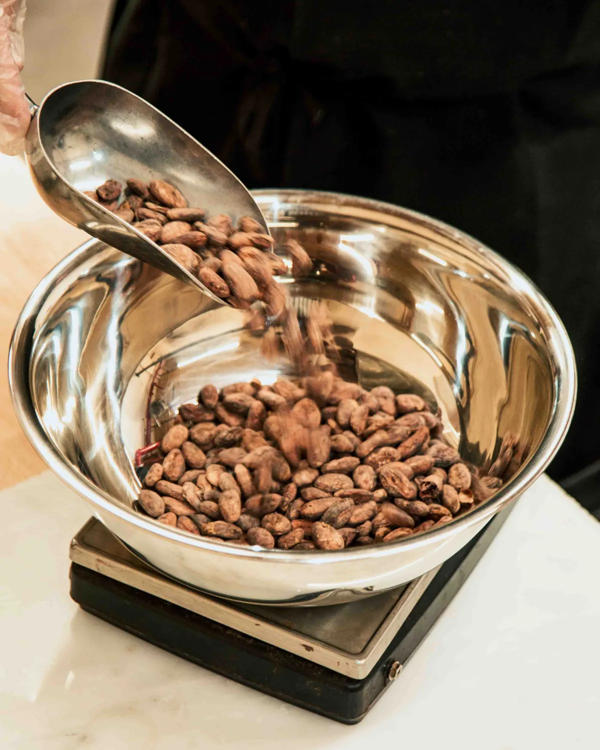 A hand pours cacao beans from a scoop into a metal bowl on a scale during a chocolate-making class.