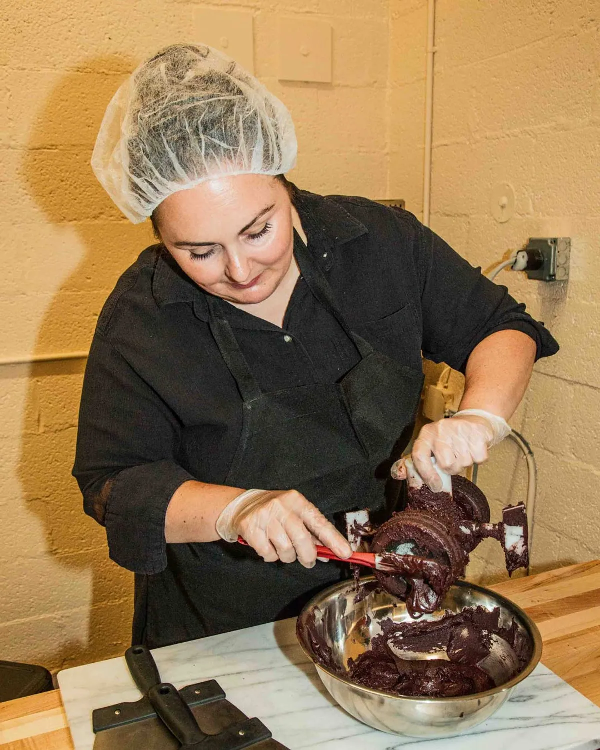 A woman in a hairnet and apron pours chocolate mixture into a bowl during a Bean-to-Bar chocolate class in NYC.