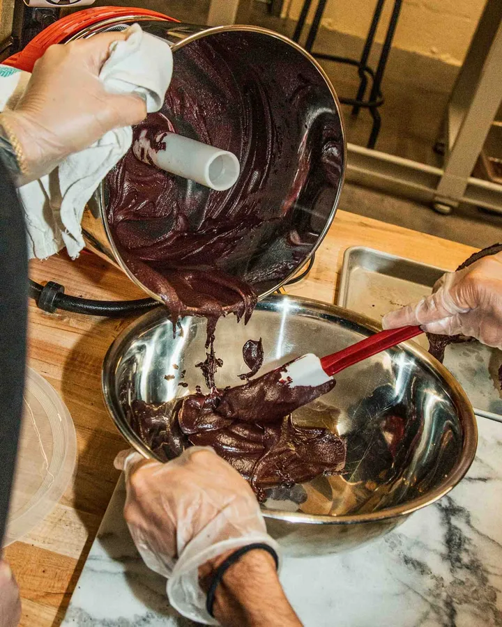 Hands in gloves mixing chocolate batter from a container into a metal bowl during a chocolate-making class.