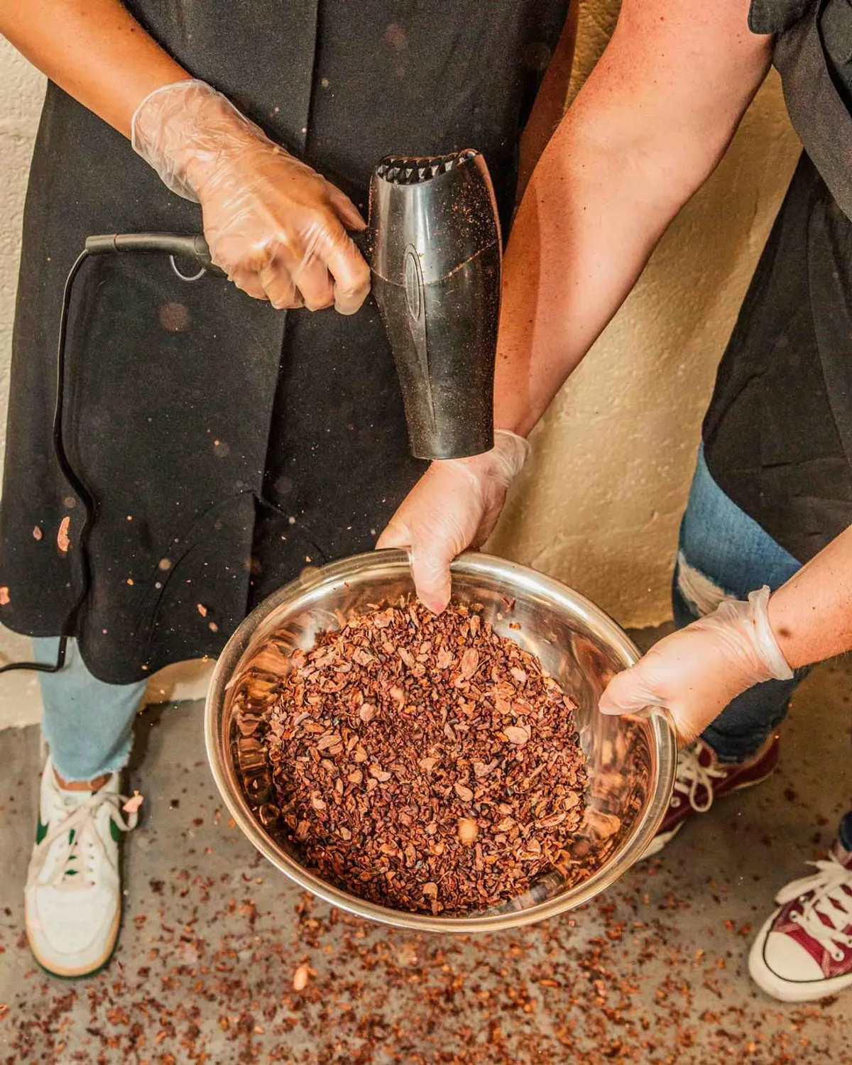 Two participants use a hairdryer to clean cocoa nibs in a metal bowl during a Bean-to-Bar Chocolate Class.