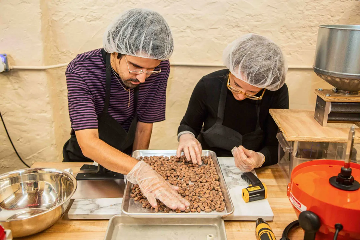 Two individuals in hairnets and gloves examine cocoa beans during a Bean-to-Bar Chocolate Class in NYC.