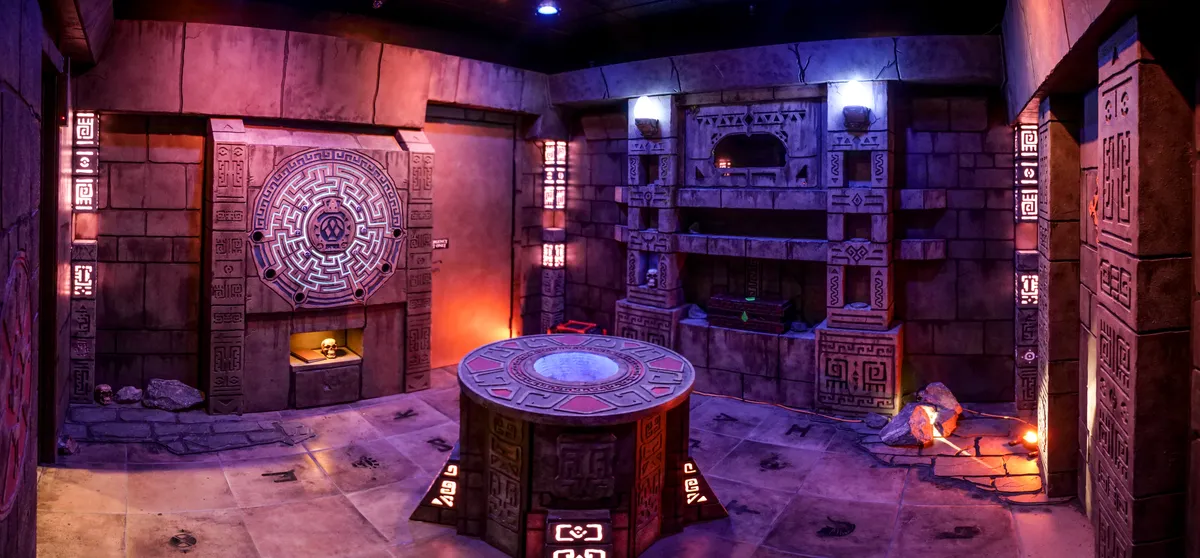 A dimly lit escape room featuring intricate stone carvings, a circular table, and a maze-like wall design.
