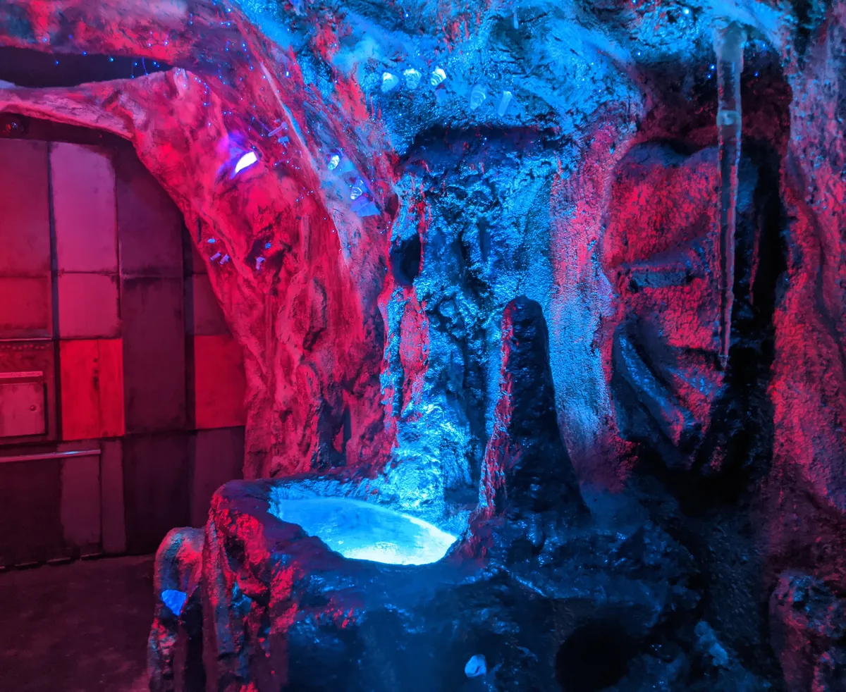 Brightly lit escape room with rocky walls in blue and red tones, featuring a small water feature.