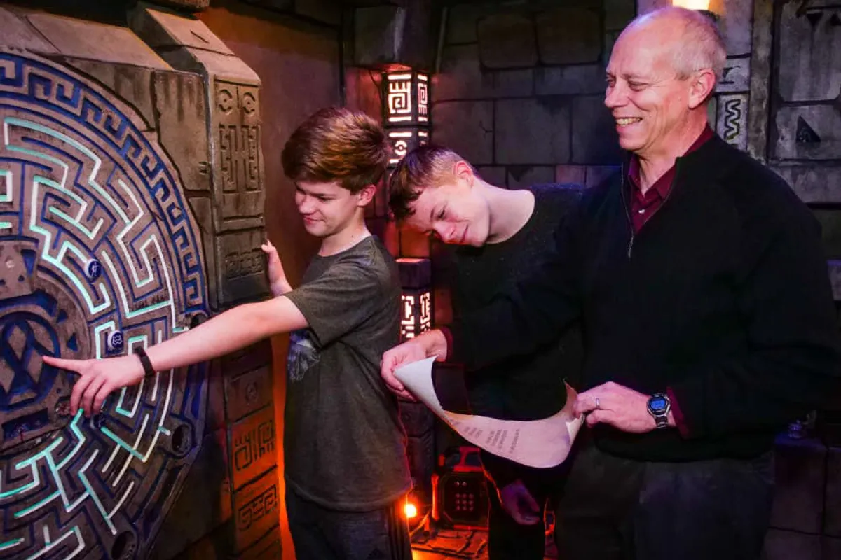 Three participants engage in a team-building escape room activity, examining a puzzle and instructions while smiling.