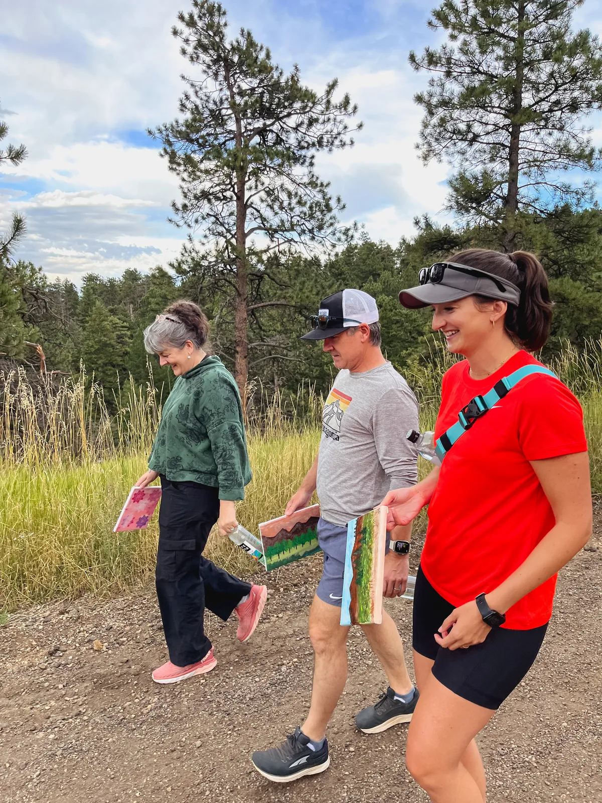 Three participants walk along a trail in Boulder, holding paintings, enjoying a team-building hike and paint activity.