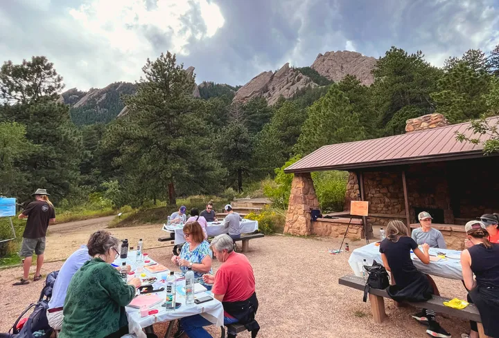 Group of people painting outdoors at a picnic area, surrounded by trees and mountains, during a team-building event.