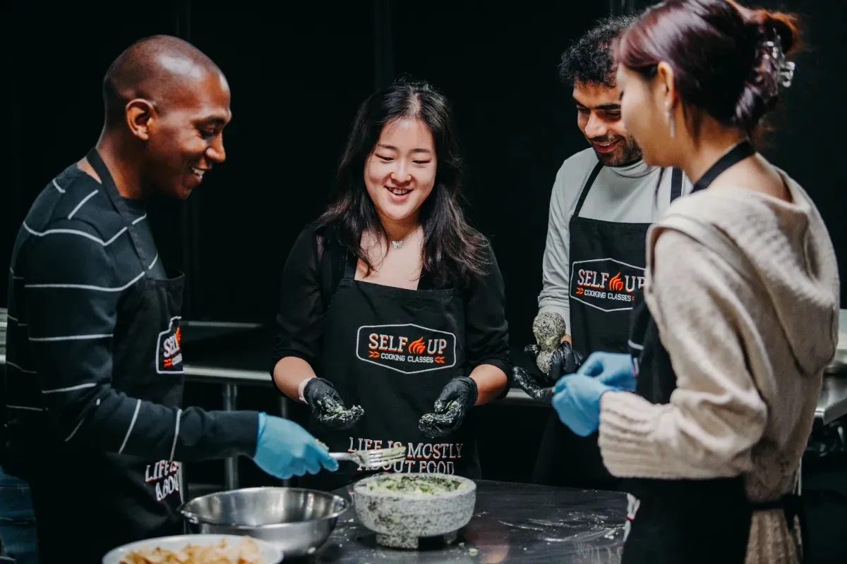 Four people in aprons engage in a Healthy Cooking Class, smiling and preparing food together in Boston.