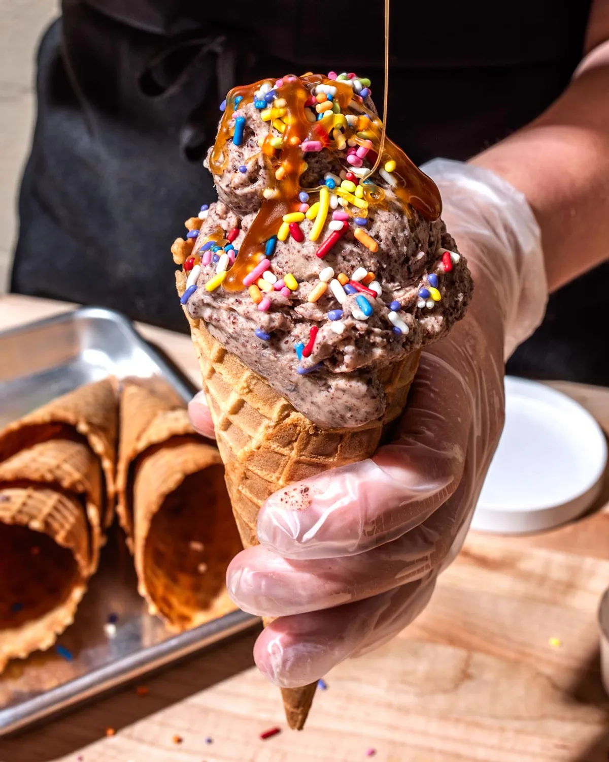Hand holding a waffle cone filled with chocolate ice cream, topped with colorful sprinkles and caramel drizzle.