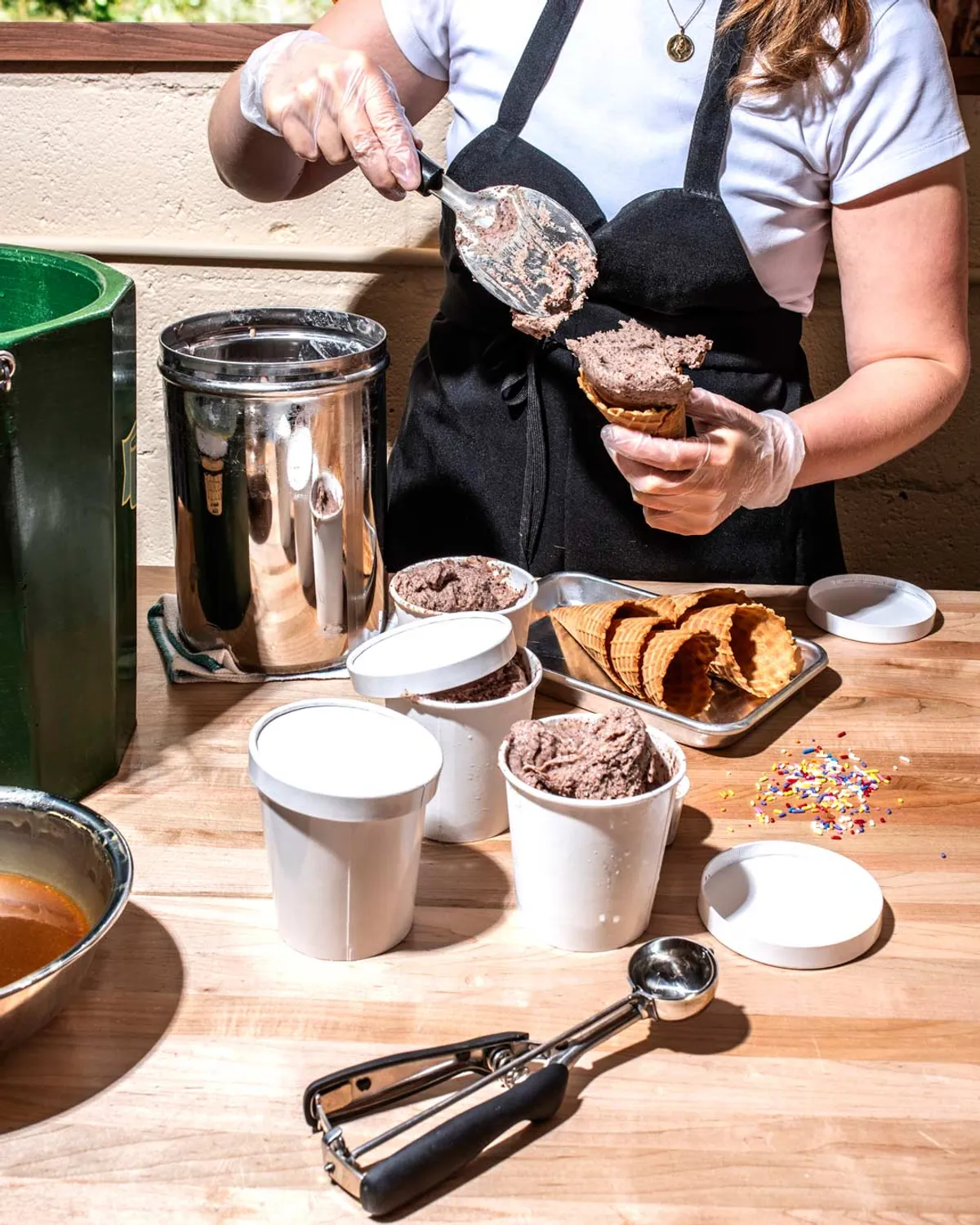 A person scoops chocolate ice cream into a cone, surrounded by ice cream tubs, cones, and sprinkles on a table.