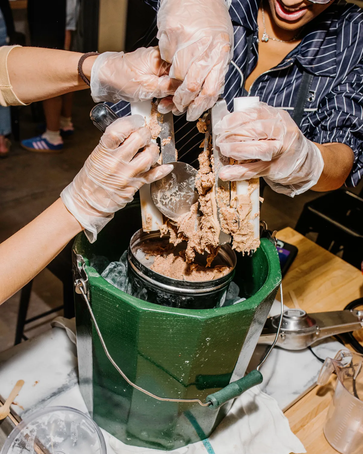 Two people in gloves extracting homemade ice cream from a churn inside a green bucket during an ice cream-making activity.