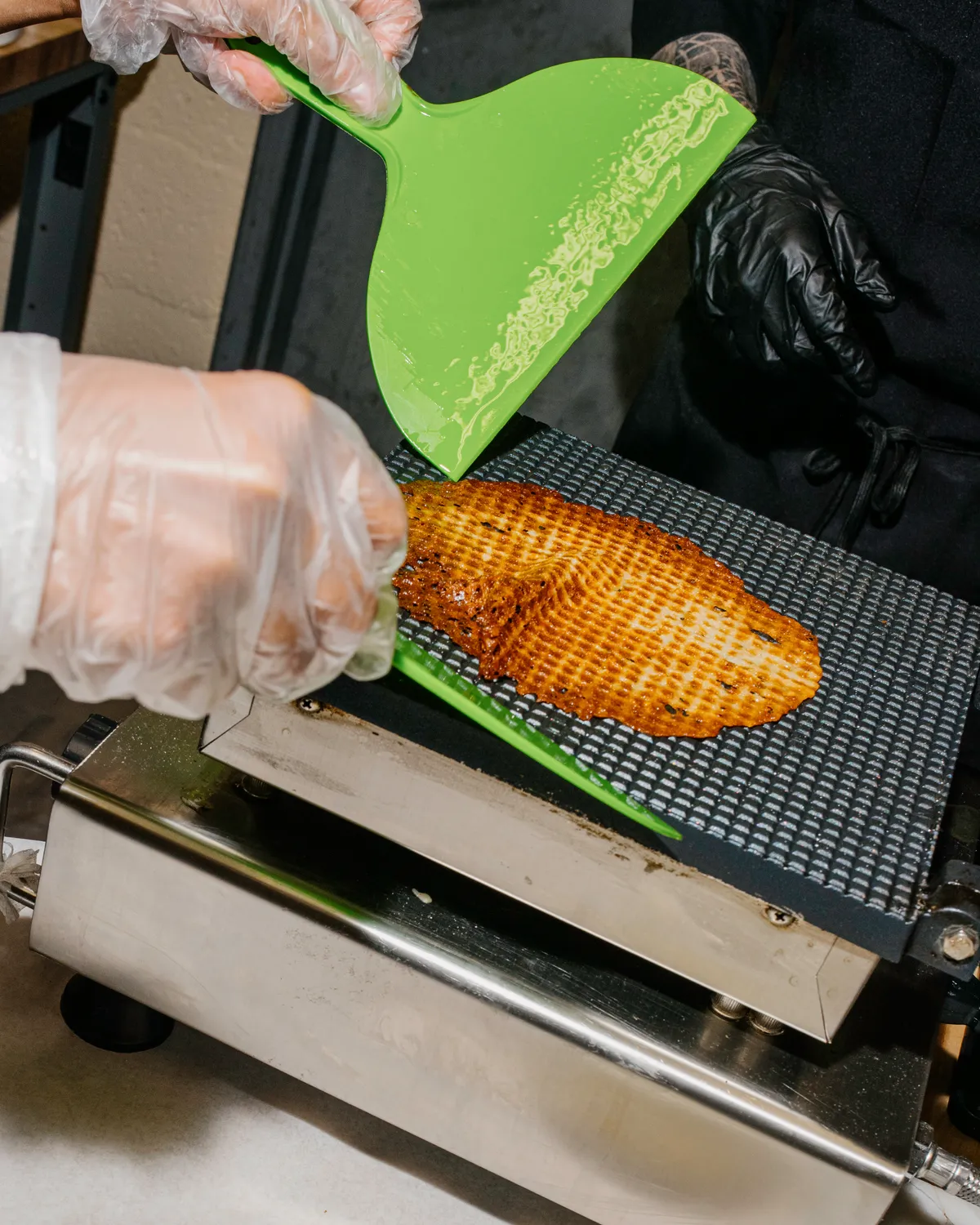 Hands in gloves using a green spatula to flip a cooked waffle on a grill during an ice cream-making activity.