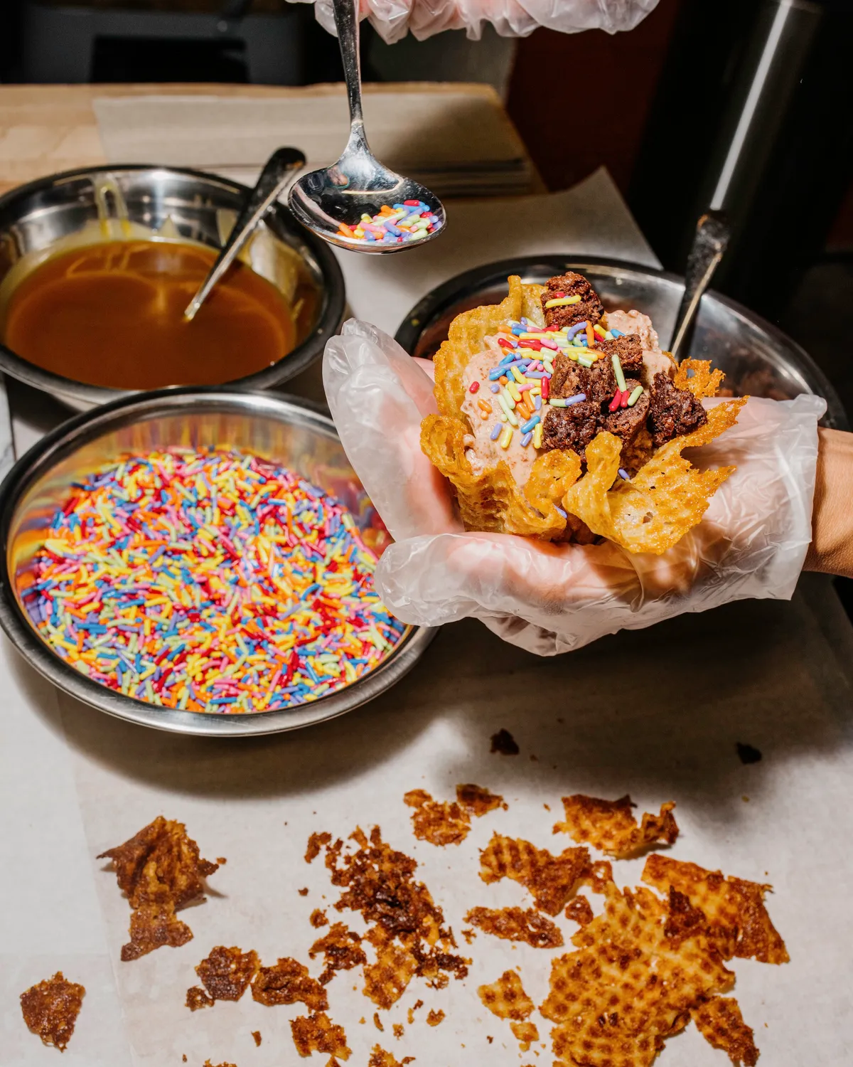 A person adds colorful sprinkles to a freshly made ice cream cone, surrounded by toppings and broken waffle pieces.