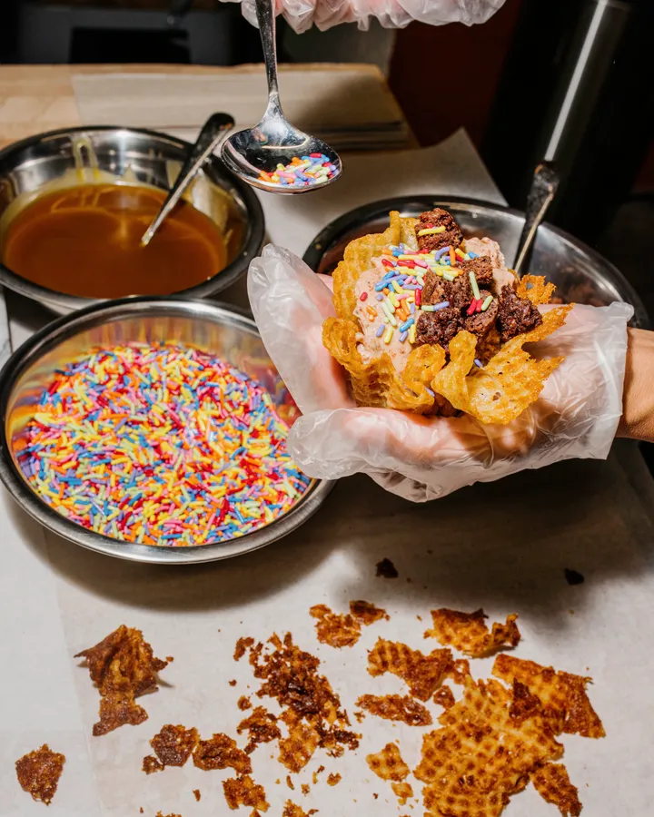 A person adds colorful sprinkles to a freshly made ice cream cone, surrounded by toppings and broken waffle pieces.