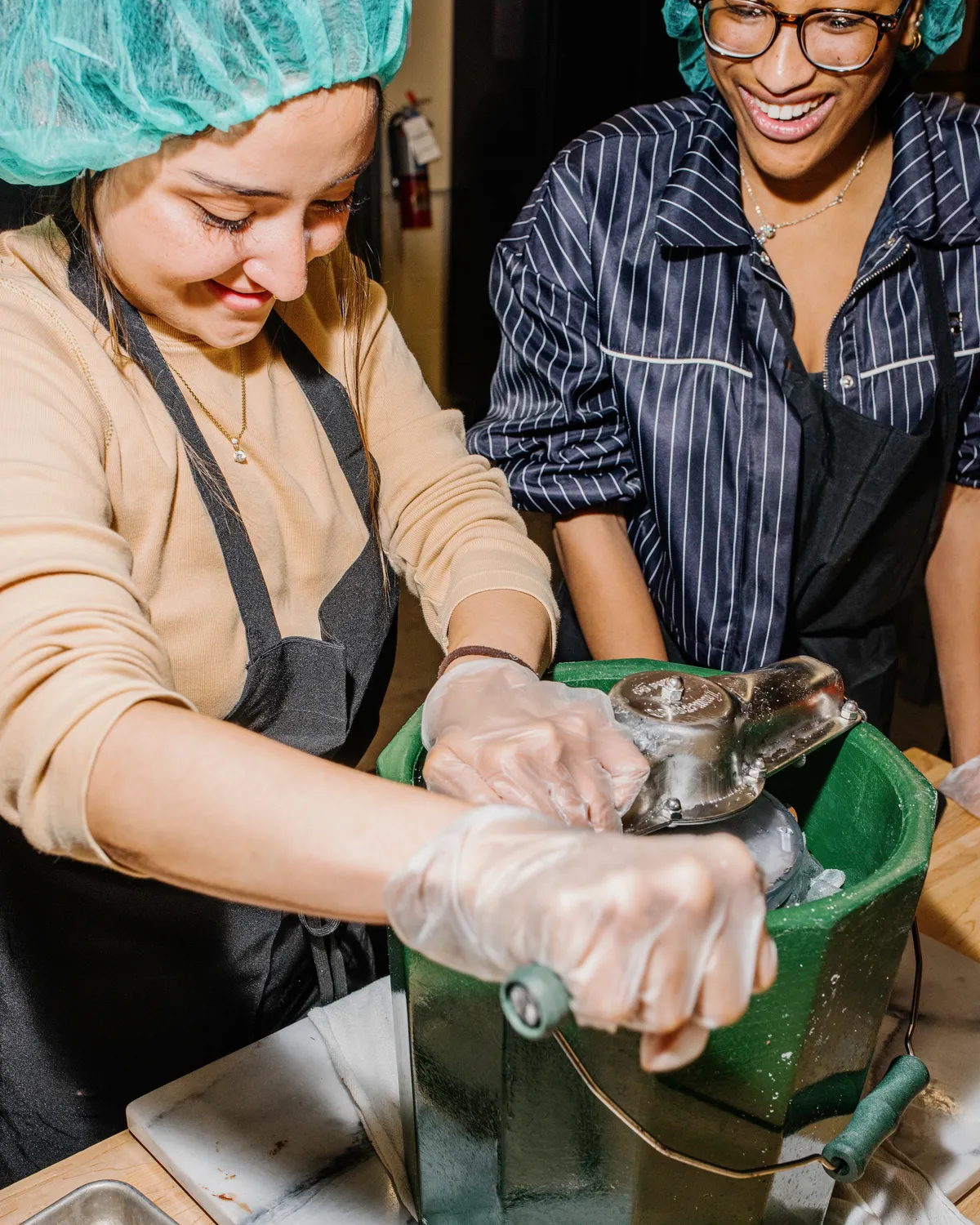 Two people making ice cream in NYC, one cranking the machine while the other smiles, both wearing aprons and gloves.