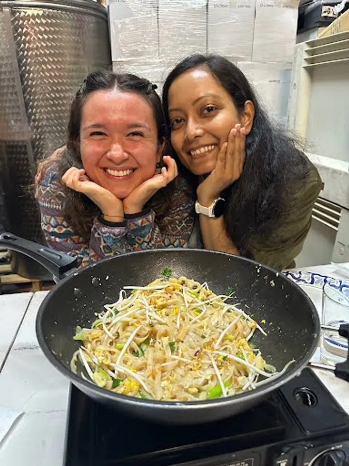 Two smiling women pose with hands on cheeks, showcasing a large pan of cooked noodles with vegetables in a kitchen setting.