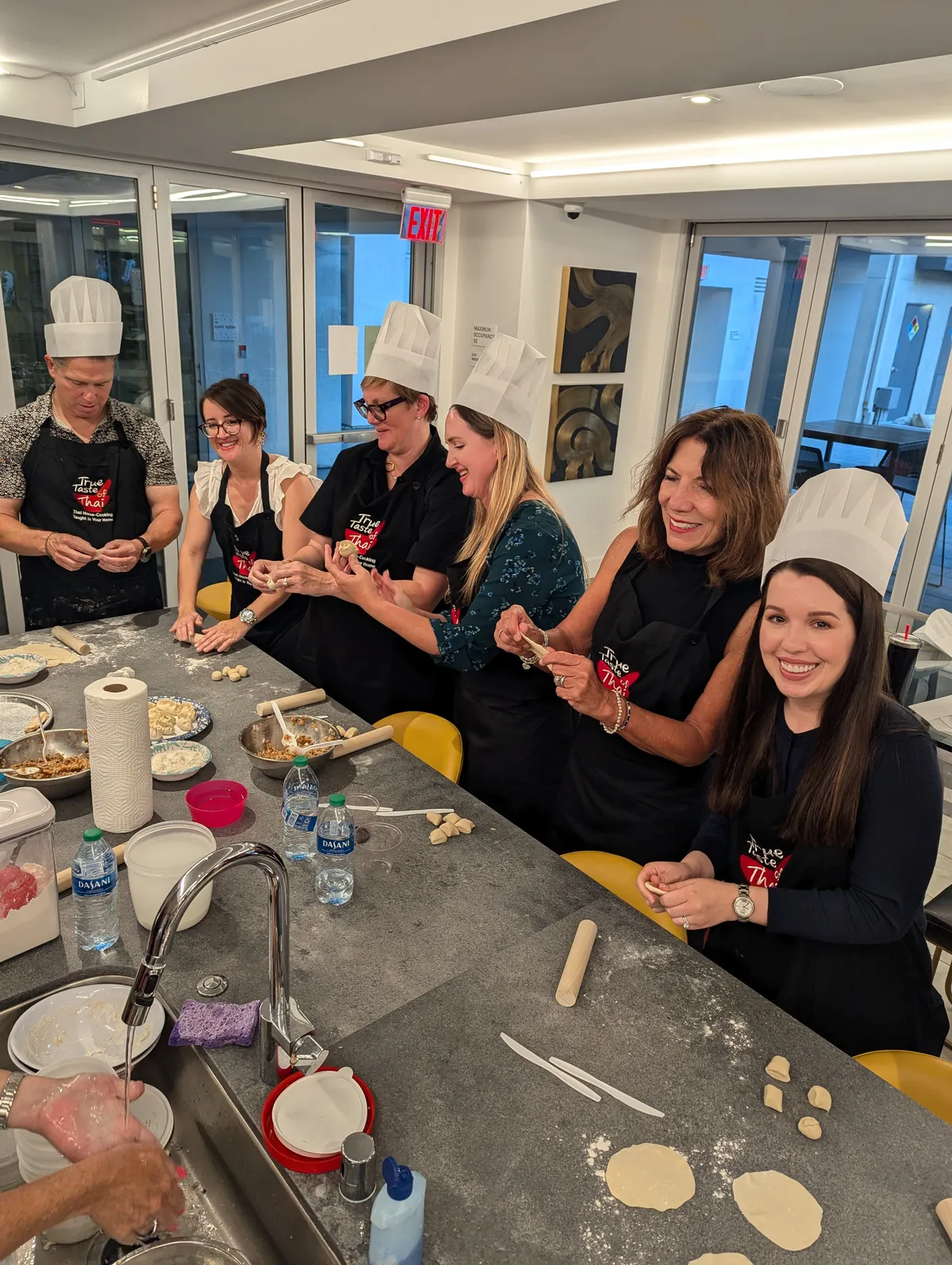 Group of six people in chef hats making gyoza dumplings at a cooking class in San Diego. Flour and ingredients on the table.