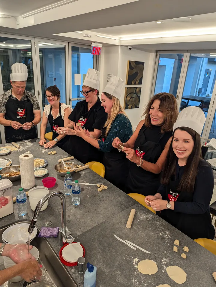 Group of six people in chef hats making gyoza dumplings at a cooking class in San Diego. Flour and ingredients on the table.