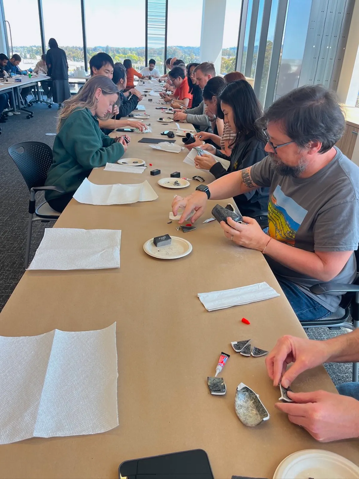 A group of people engaged in a Kintsugi class, repairing pottery at a long table in a bright room with large windows.