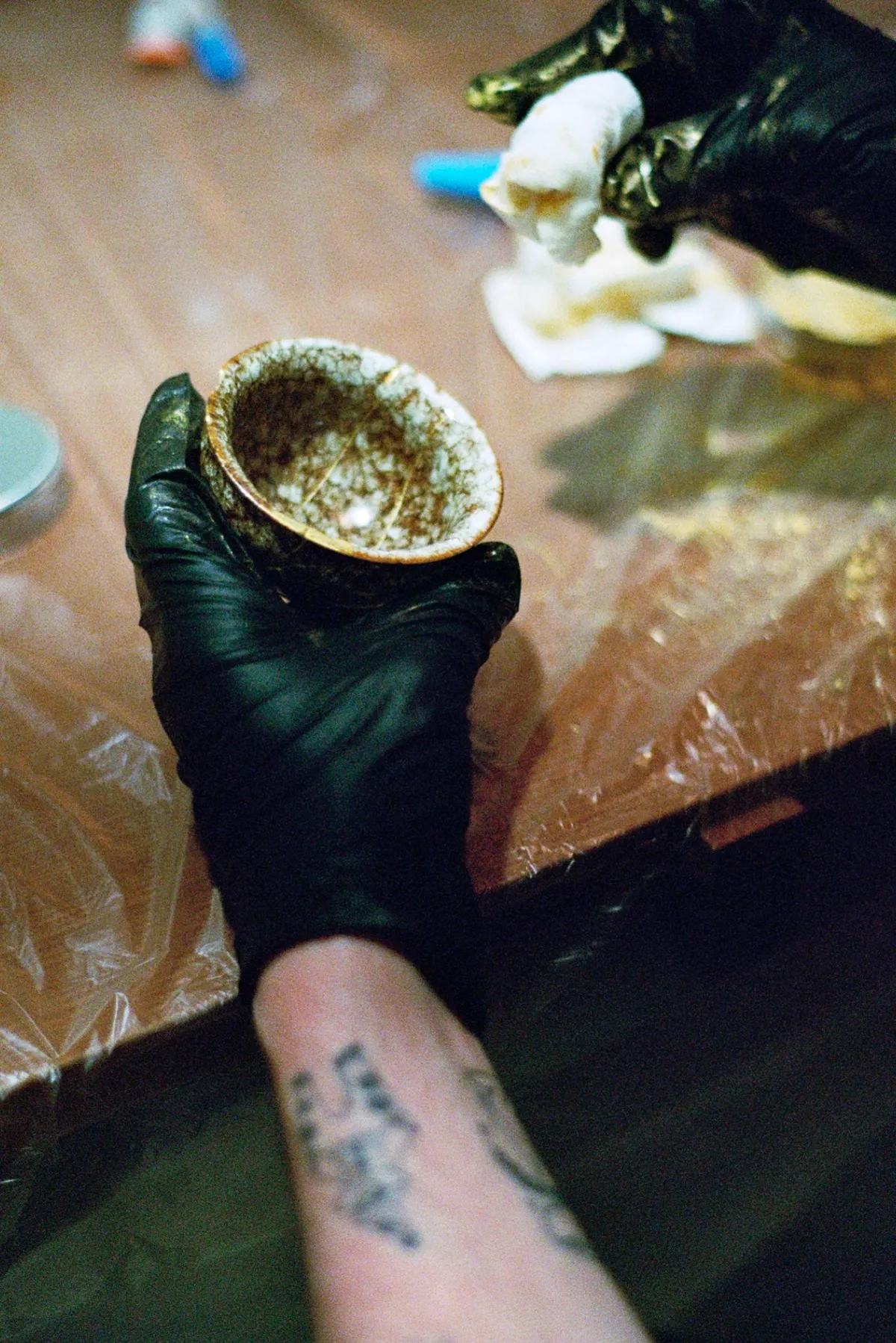 A person wearing black gloves holds a bowl, working on a Kintsugi project at a table covered with plastic.