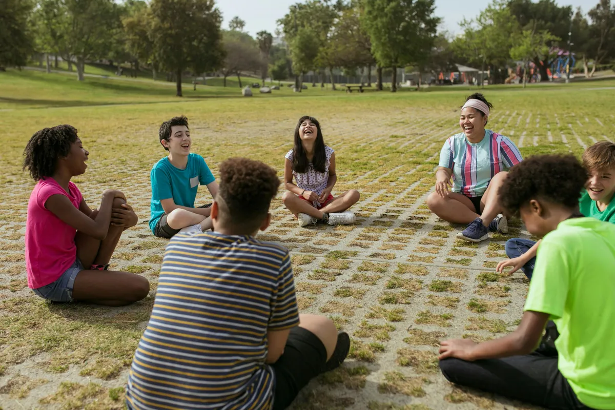 A group of children sit in a circle on grass, laughing and enjoying a meditation class in a park.