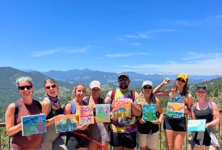 A group of eight people holding their painted artworks against a scenic mountain backdrop during a team-building hike.