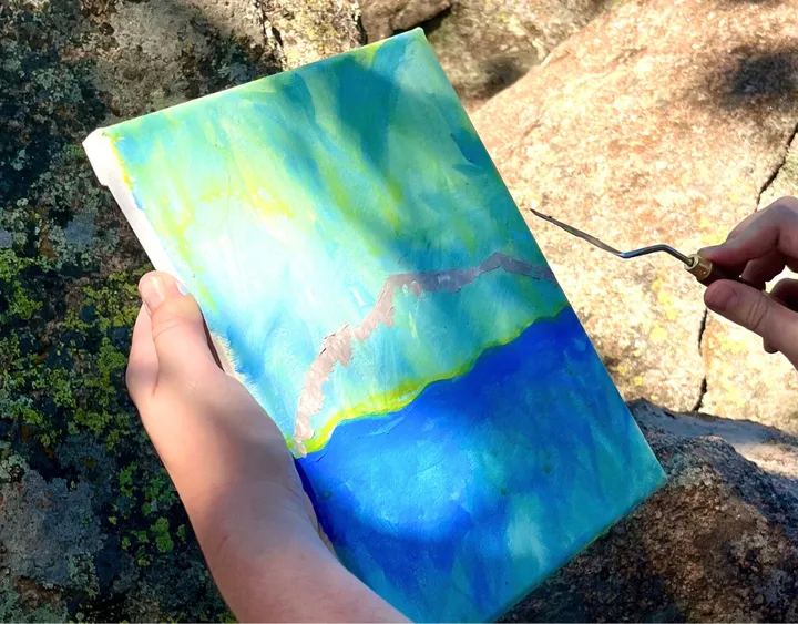A hand holds a colorful canvas with blue and green paint, set against rocky outdoor scenery.