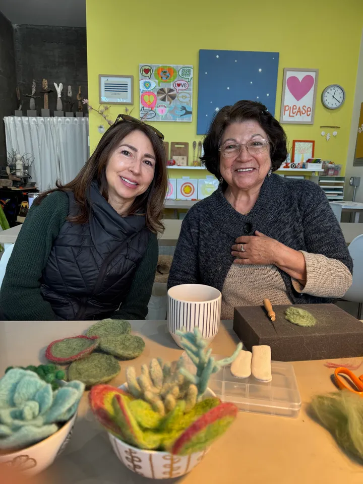 Two women smile in a craft studio, surrounded by needle-felted succulents and tools, promoting a creative activity.