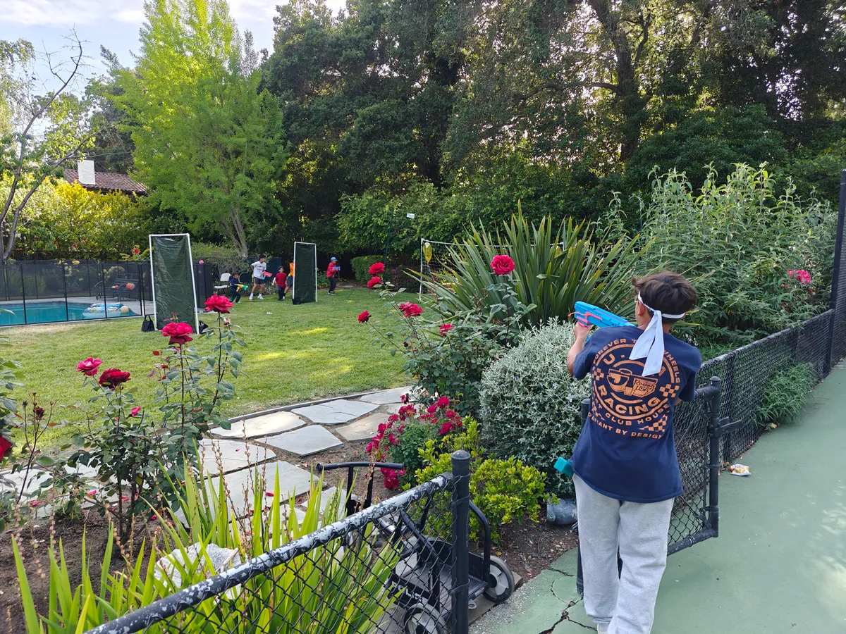 A person with a Nerf blaster stands near roses, while others participate in a Nerf Battle team-building activity.