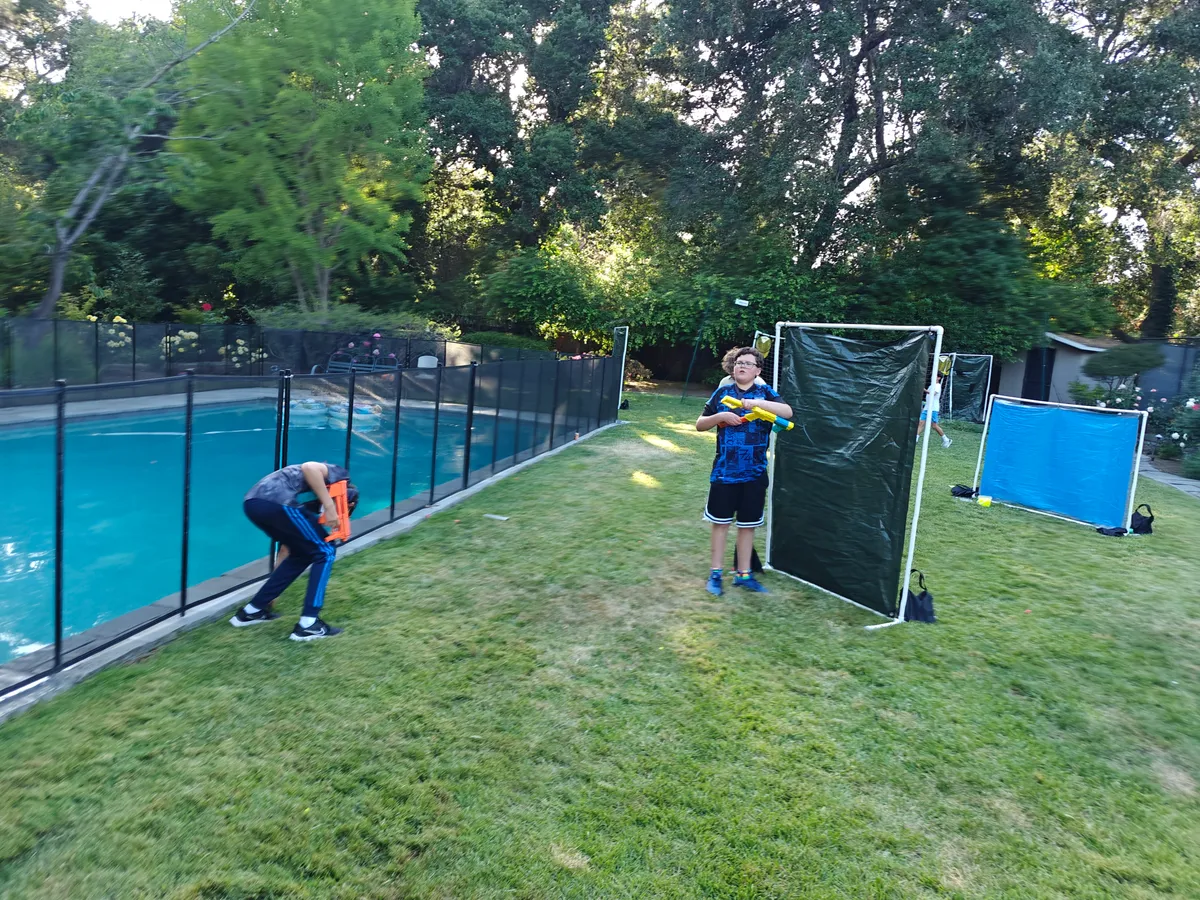 Two kids play Nerf Battle in a grassy area by a pool, using barriers for cover. Bright sunshine and greenery surround them.