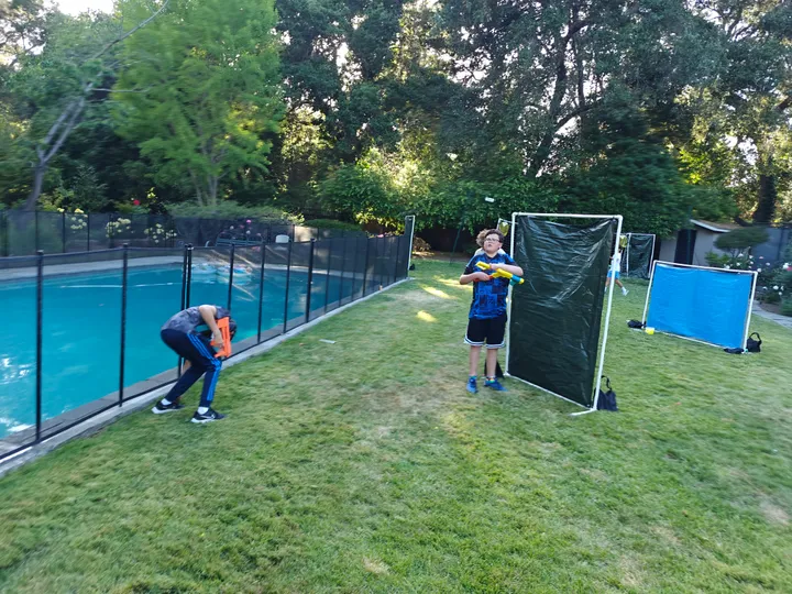 Two kids play Nerf Battle in a grassy area by a pool, using barriers for cover. Bright sunshine and greenery surround them.