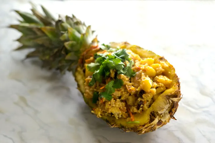 A pineapple half filled with fried rice, garnished with cilantro, set on a marble surface.