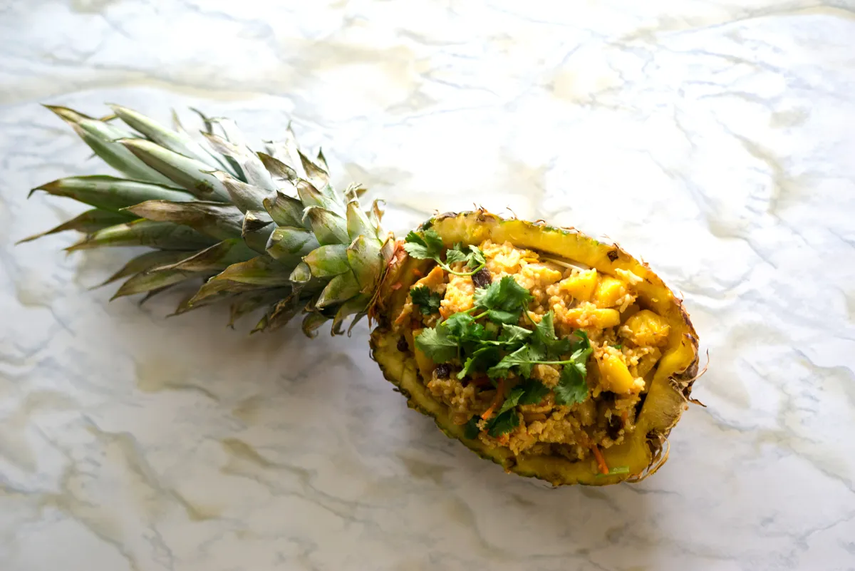 Pineapple half filled with fried rice, topped with cilantro, on a marbled surface. Ideal for a team-building activity.