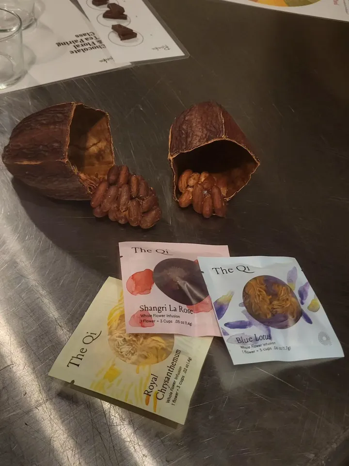 Cocoa pods with beans beside three tea packets: Royal Chrysanthemum, Shangri La Rose, and Blue Lotus on a table.