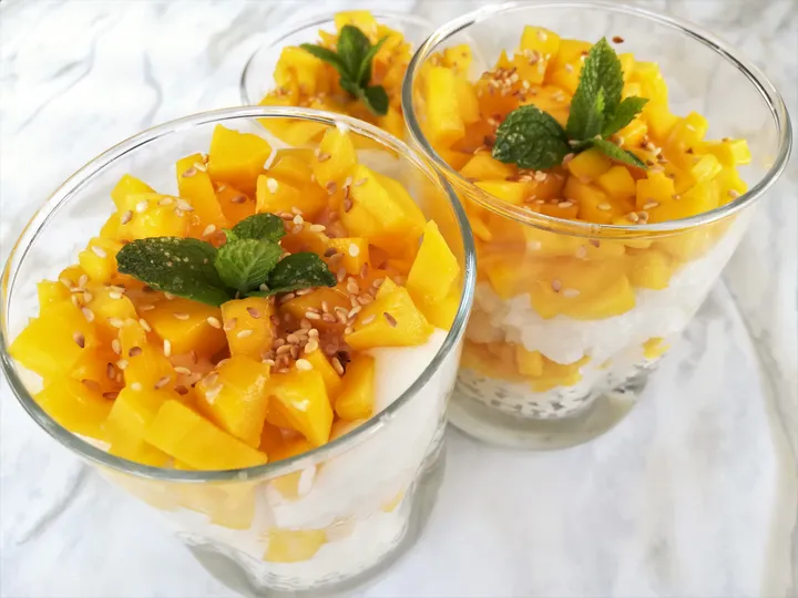 Two glasses of mango sticky rice topped with fresh mango, mint leaves, and sesame seeds on a marble surface.