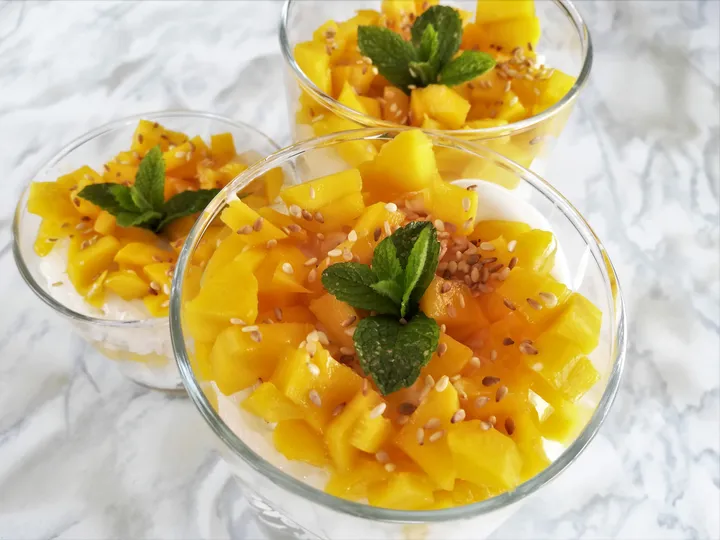 Three cups of mango sticky rice topped with fresh mango, sesame seeds, and mint leaves against a marble background.