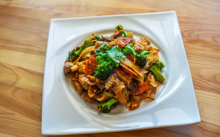A white plate holds a colorful Thai Pad-See-Ew dish with vegetables and cilantro, resting on a wooden surface.