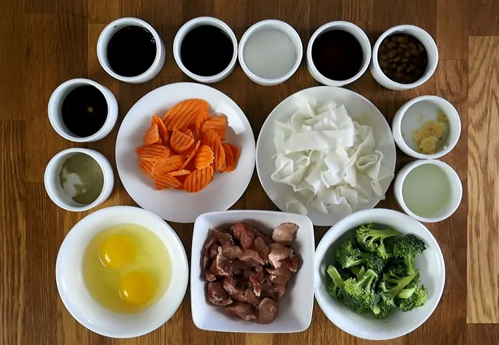 Ingredients laid out for a Thai Pad-See-Ew class: vegetables, sauces, noodles, eggs, and meat on a wooden surface.