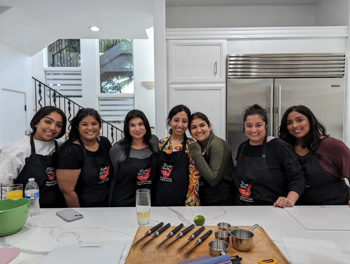 Group of seven women in black aprons smiling in a kitchen during a Thai cooking class in San Diego.