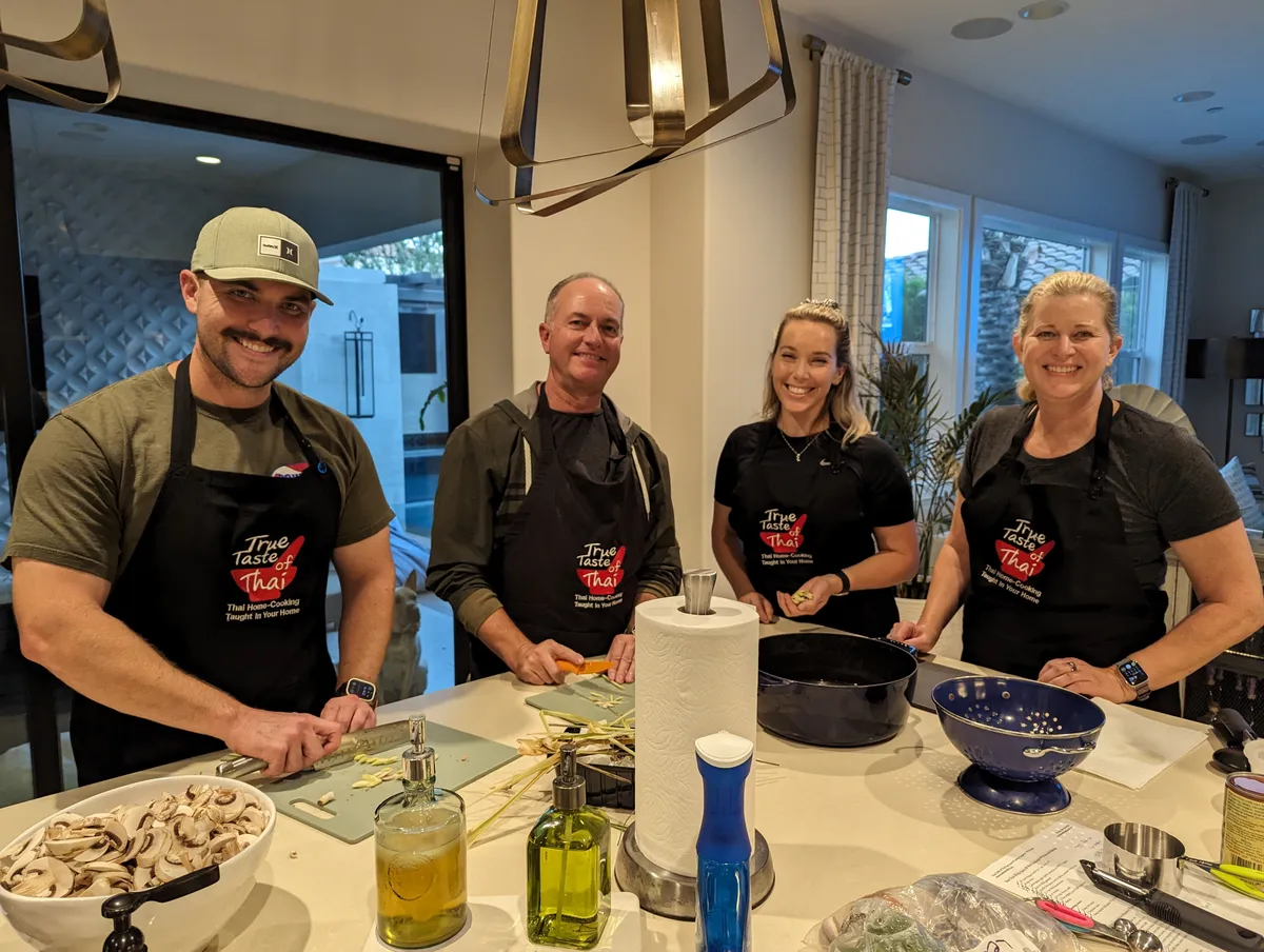Four smiling participants in black aprons prepare ingredients for Pad-See-Ew in a bright kitchen.