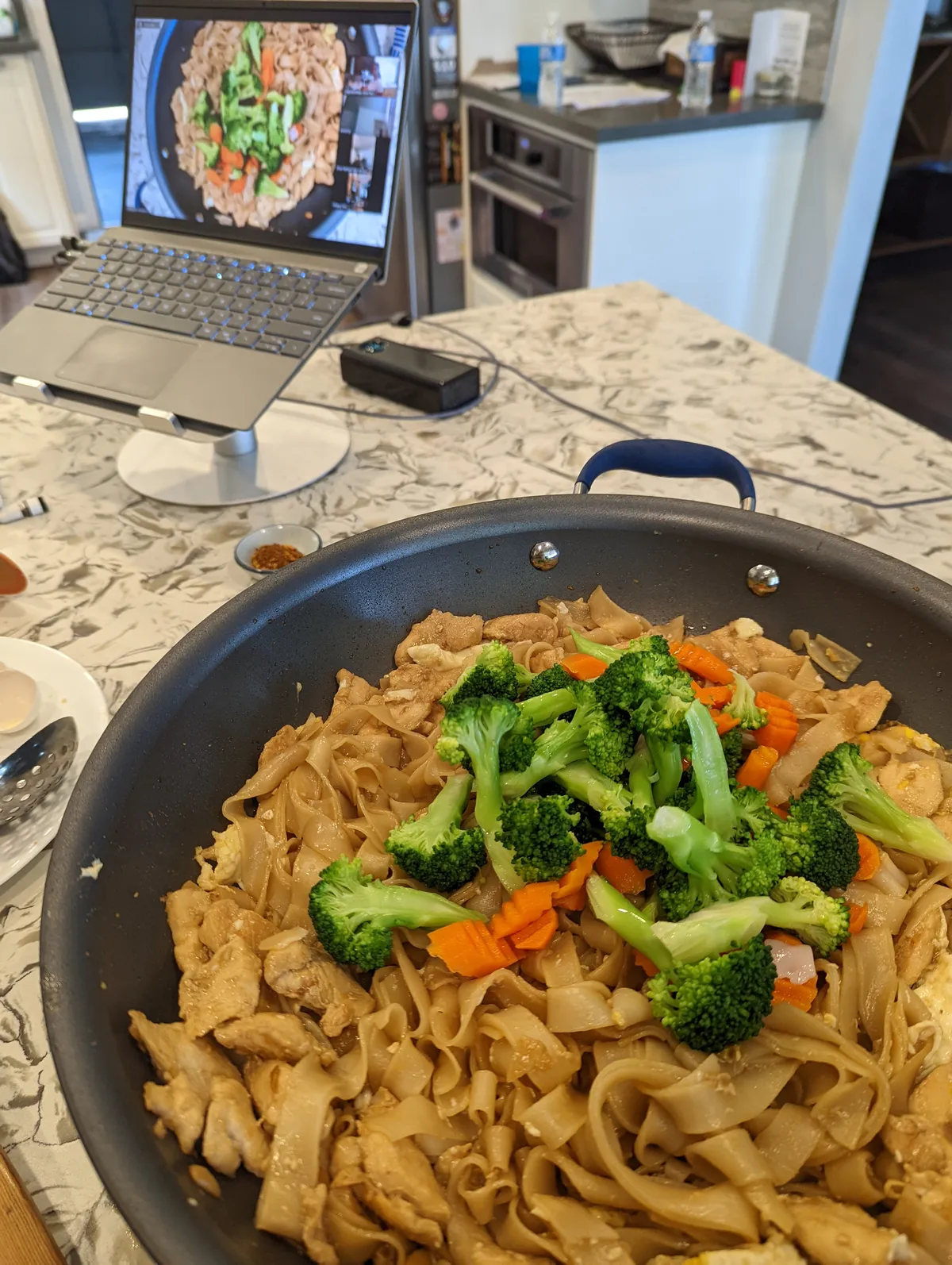 A pan of Pad-See-Ew with broccoli and carrots, beside a laptop displaying a cooking class in San Diego.