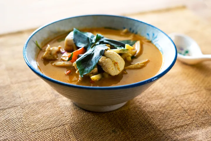 A bowl of Thai red curry with chicken, vegetables, and fresh basil, placed on a textured table surface.