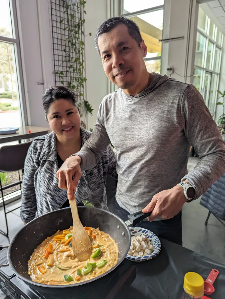 Two people in a cooking class stir Thai red curry in a large pan, with vegetables and tofu on the table.